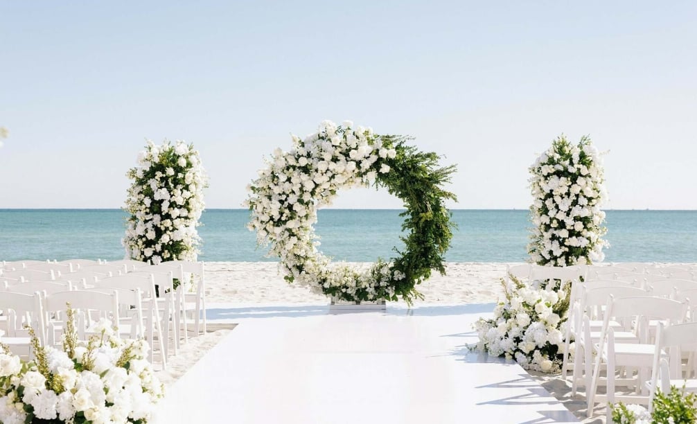 A beach wedding setup with a white archway, flowers, and chairs facing the ocean.